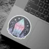 A circular sticker design featuring a pastel iridescent crystal surrounded by mystical foliage and twinkling stars on a dark background.