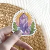 A vibrant sticker depicting a collection of amethyst crystals in shades of purple and clear, surrounded by green leaves and a glowing yellow-orange aura.