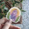 A vibrant, hand-drawn sticker depicting a cluster of purple and light blue crystals with green leafy accents, set against a radiant yellow sunburst.