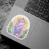 A vibrant sticker featuring a cluster of purple and blue crystals emanating a soft glow, surrounded by green leaves, with a pastel sunburst background.