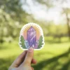 A vibrant sticker featuring a cluster of purple crystals emitting a soft glow, surrounded by green leaves against a blurred natural background.