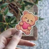This adorable, blushing bear clutching a pink heart is a waterproof vinyl sticker, perfect as a UV resistant decal for your water bottle or laptop.