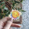 A cheerful cartoon sun with a rainbow halo and sparkling accents, held by a hand.