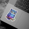 A cute cartoon owl with purple and blue starry feathers and large golden eyes perches on a brown branch with green leaves.