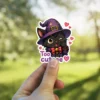 An adorable winking black cat wearing a purple witch hat and a red bow, surrounded by hearts with the text 'Too cute'.
