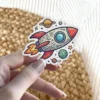 A colorful cartoon rocket ship sticker with doodle art on its body, flying past planets and stars on a white background.