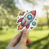 A cartoon rocket ship sticker with fun doodle patterns, blasting through space surrounded by planets and stars.