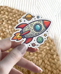 A colorful cartoon rocket ship sticker with doodle art on its body, flying past planets and stars on a white background.