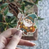 A friendly cartoon gnome with a white beard, green hat, and tool belt walks while carrying an axe and a sack of carrots.