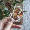 A detailed illustration of a cute baby dragon with golden scales and orange wings, sitting and looking forward with a mischievous expression. The sticker has a white border.