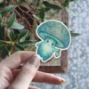 A charming aqua and teal mushroom character sticker with a smiling face, intricate swirling patterns on its cap and body, holding a small glowing lantern.