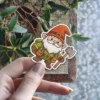 A friendly cartoon garden gnome with a white beard, red hat, and green tunic holds a small axe.
