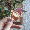 A delightful cartoon gnome sticker featuring a white beard, purple hat, yellow shirt, orange overalls, and a friendly smile.