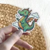 A cute green cartoon dragon with orange wings happily flying among fluffy white clouds on a die-cut vinyl sticker.