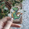 A playful, smiling green dragon with orange scales and wings, surrounded by fluffy white clouds in a cartoon style sticker.
