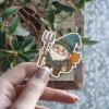 A charming cartoon gnome character sticker, wearing a blue hat and tunic, holding a pitchfork, with autumn leaves in a backpack.