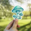 A whimsical sticker of three cute blue-green mushrooms; the central one smiles, surrounded by two smaller companions with detailed caps.