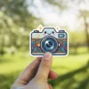 A kawaii cartoon camera sticker with a smiling face and a floral pattern on the body.