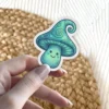 A kawaii teal mushroom sticker with a happy face and swirling patterns on its cap is held between fingers.