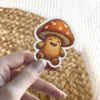 A cheerful, hand-drawn style sticker of a happy mushroom character with a textured, knitted brown body and an orange cap with white spots.