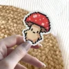 A charming pixel art style mushroom sticker with a red cap and a light brown stem, featuring a simple, friendly face.