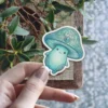 A charming teal mushroom character with a happy face and an ornate, jewel-topped cap is shown against a natural background.