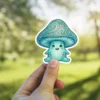 A cute teal mushroom character with a smiling face and swirling patterns on its cap is held up against a blurred green background.