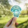 A cheerful, light blue mushroom character sticker featuring intricate mandala patterns on its cap and base, holding a small apple.