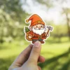 A cheerful cartoon gnome sticker with an orange hat, white beard, holding a silver axe and carrying a sack with a small tree.