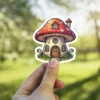 A charming sticker depicts a whimsical mushroom house with a red spotted cap, windows, a wooden door, and small flowers.