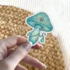 A whimsical teal mushroom character with a decorative cap and a glowing lantern is held by a person's fingers.
