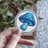 A circular sticker features a charming blue mushroom with a smiling face, adorned with glowing runes.