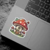 A whimsical sticker depicts a large red mushroom house with white polka dots and a small green bug on top, surrounded by smaller mushrooms and grass.