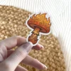 A sticker featuring a mushroom with a cap engulfed in flames and a stem decorated with intricate patterns, all rendered in vibrant orange and yellow hues.