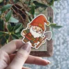 A smiling cartoon garden gnome with a red hat and white beard holds a small shovel.