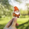 A cute cartoon sticker of a friendly garden gnome with a white beard carrying a backpack with a plant sapling.