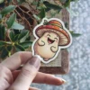 A whimsical sticker features a smiling mushroom character wearing a spotted hat, held by a person's hand.