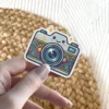 A cute light blue camera sticker with a smiling face and a colorful floral pattern around the lens.