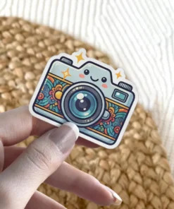 A cute light blue camera sticker with a smiling face and a colorful floral pattern around the lens.