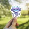 A kawaii cartoon jellyfish sticker with a smiling face and trailing tentacles is held between fingers.