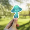 A detailed teal mushroom sticker with intricate swirling patterns, held by a person's fingertips against a blurred green background.