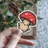 A pixel art mushroom sticker with a red cap and beige stem is being held by a hand.