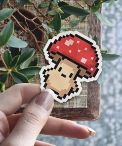 A pixel art mushroom sticker with a red cap and beige stem is being held by a hand.