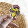 A yellow mushroom with black stripes, a pink mohawk, and a mischievous expression is depicted as a vinyl sticker.