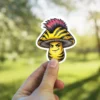 A vinyl sticker of a cartoon mushroom with a yellow and black striped cap and a mohawk hairstyle. The mushroom has a mischievous expression.