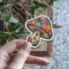A vinyl sticker featuring a whimsical mushroom design pieced together from colorful quilt squares and adorned with buttons.