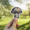 A sticker of a mushroom designed to look like stained glass, held by a person's fingers.