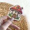 A whimsical cartoon sticker features a charming mushroom house with a red spotted cap, wooden door, and small windows, surrounded by grass and tiny flowers.