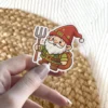 A cute cartoon garden gnome with a white beard, red hat, and green tunic holds a pitchfork and carries a sack on his back.