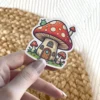 A cute sticker of a mushroom house with a red and white spotted cap, a friendly green caterpillar on top, and smaller mushrooms around it.
